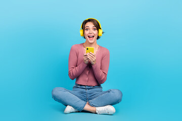 Photo of astonished crazy girl sit floor use smart phone listen music isolated on blue color background