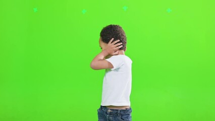 Naughty african american little boy covering ears from noise, twisting inside. Front view of funny male kid in t-shirt keeping hands on ears, isolated on green studio background. Concept of gesturing.