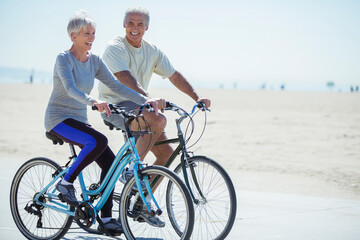 Senior couple riding bicycles on beach boardwalk - obrazy, fototapety, plakaty