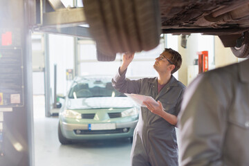 Mechanic with clipboard under car in auto repair shop - obrazy, fototapety, plakaty