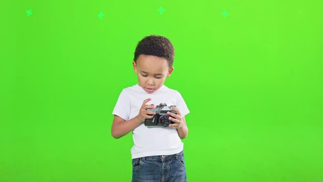 Focused little boy in t-shirt holding, examining retro vintage photo camera in studio. Front view of pretty african american child taking photo, isolated on green background. Lifestyle, hobby concept.