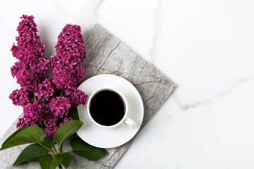 Branch of burgundy lilac flowers and cup of fresh black coffee on white background with a marble texture with a place for text. Flat lay, copy space