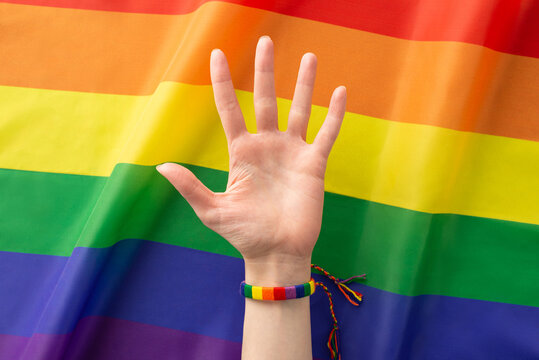 Top-down photo of a non-binary person's hand, adorned with a rainbow bracelet and fingers spread wide, against a wavy LGBTQ flag background