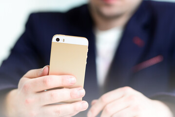 Young businessman working on the gold smartphone at work in blue suit in interior behind the office desk.