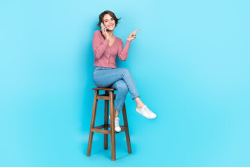 Full length photo of positive girl wear pink cardigan sit on chair talk on phone directing empty space isolated on blue color background
