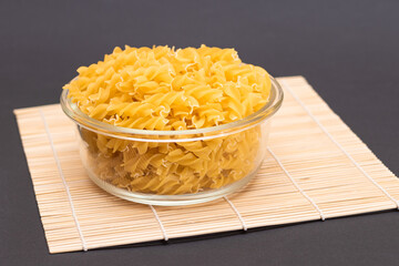 Uncooked Fusilli Pasta in Glass Jar on Bamboo Mat on Black Background. Fat and Unhealthy Food. Classic Dry Spiral Macaroni. Italian Culture and Cuisine. Raw Pasta
