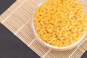Uncooked Chifferi Rigati Pasta in Glass Jar on Bamboo Mat on Black Background. Fat and Unhealthy Food. Classic Dry Macaroni. Italian Culture and Cuisine. Raw Pasta