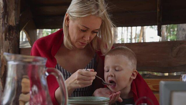 A young blonde woman feeds her little son with soup during lunch. A hungry child receives care and attention from his parent. Mom and baby eat together in a cafe.
