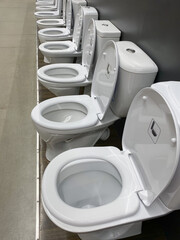 Exhibition of samples of сeramic white toilet bowls standing in a row in the warehouse of a plumbing store. Samples of modern sanitary ware for the toilet. New modern toilet in the plumbing store