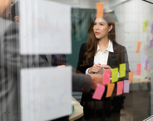 Businessperson teamwork meeting in office and use sticky post notes to brainstorming ideas on glass board. Asian businesswoman planning corporate strategy with colleague for success startup business.