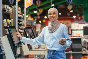 Self-service in a supermarket. A young Arab woman in a hijab is standing near the electronic cash...