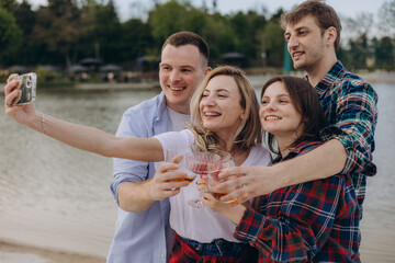 Obraz premium Stylish young people in casual clothes waving hand, posing for smartphone camera when taking selfie or shooting video for social media using mobile phone by lake. Friends having rest and fun.