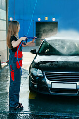 Woman Worker in Uniform Washing Car with High Pressure Gun with Hose at Car Wash Station.