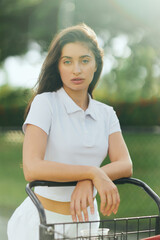 Obraz premium tennis court in Miami, sporty young woman with brunette hair standing in white polo shirt near tennis cart, blurred green background, looking at camera, pretty tennis player, soft filter