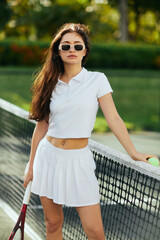 Fototapeta tennis court in Miami, athletic young woman with long hair standing in white outfit and sunglasses while holding racket and ball near tennis net, blurred background, iconic city, looking at camera obraz