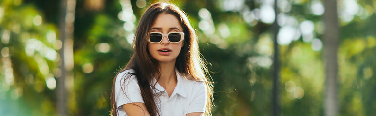 captivating young woman with long hair standing in white polo shirt and trendy sunglasses near...