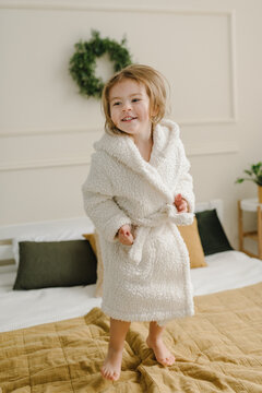 Portrait of beautiful child wearing a bathrobe, jumping on bed and smiling after shower or bathing at bedroom. Cute adorable caucasian cheerful smiling blond girl. Kid in a home bathrobe. Closeup.