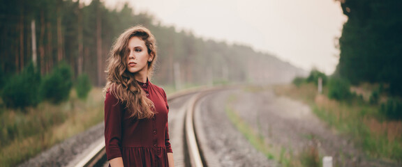 Beautiful girl with curly natural hair enjoy nature in forest on railway. Dreamer lady in burgundy dress walk on railroad. Female portrait of inspired girl on rails at dawn. Sun in hair in autumn.