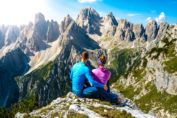 Naklejka na ściany i meble Sitting young couple enjoys epic view on Cadini di Misurina mountain range in the morning. Tre Cime, Dolomites, South Tirol, Italy, Europe.