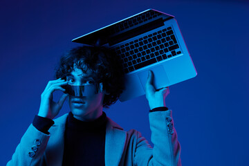 Portrait of a stylish male hacker with a laptop and futuristic glasses in blue light, cyber...