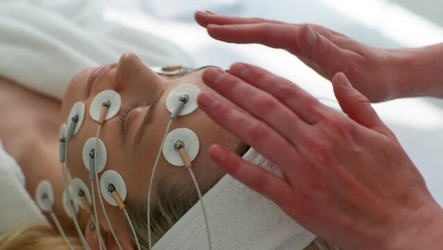 Close-up top rear view of pretty young woman patient preparing for myostimulation procedure in modern beauty clinic. Physiotherapist putting electrodes of myostimulator during bioresonance therapy.