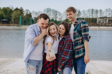 Group of stylish young people in casual clothes waving hand, posing for smartphone camera when taking selfie or shooting video for social media using mobile phone by lake. Friends having rest and fun.