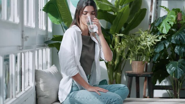 Video of young woman in lotus position drinking water while sitting on couch in the living room full of plants at home