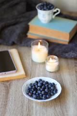 Cup of blueberries, set of books, phone, eyeglasses, lit candles and blanket on the table. Hygge at home. Selective focus.