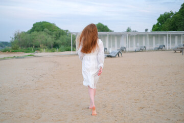 Beautiful redheaded girl on the beach in summer running on the sand in a white dress.