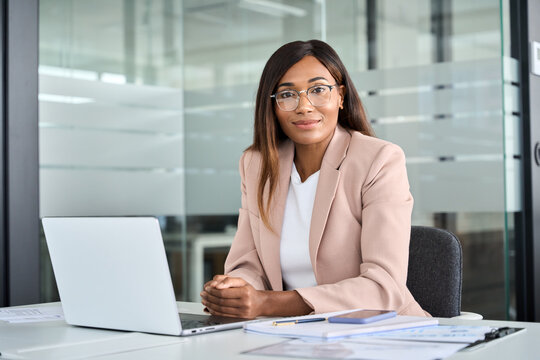 Confident Young Female Lawyer, Professional African American Business Woman Company Manager Executive Wearing Suit Glasses Working On Laptop In Office Sitting At Desk Looking At Camera, Portrait.