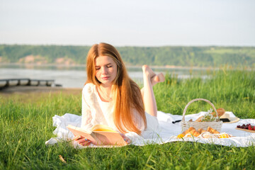 A beautiful red-haired girl in a white dress meets the summer dawn on the green grass during a picnic.
