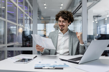 Successful businessman in casual shirt doing paperwork, boss with beard and glasses sitting at desk at workplace using laptop working with documents, holding hand up celebrating victory and triumph.