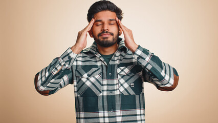 Fototapeta premium Handsome indian bearded man in shirt rubbing temples to cure headache problem, suffering from tension, migaine, stress, grimacing in pain, high blood pressure isolated alone on beige wall background