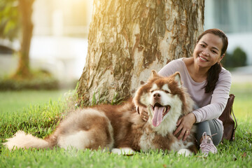 Portrait of happy Vietnamese woman patting samoyed dog when sitting under tree in park
