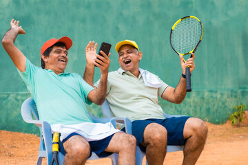 Happy laughing indian senior friends at tennis court by watching mobile phone - concept of taking break, social media and technology