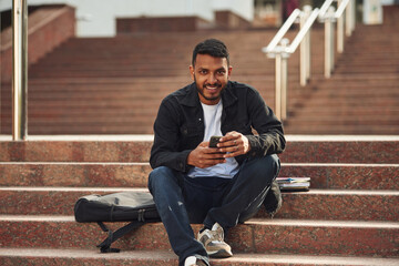 Handsome Indian student is outdoors sitting on the stairs with smartphone
