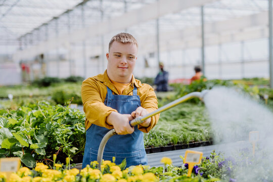 Happy Young Employee With Down Syndrome Working In Garden Centre, Watering Plants With A Shower Head And Hose.