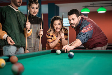 Fototapeta premium Group of friends playing billiards in a game room - Bearded young player preparing to strike the ball with cue while teammate gives instructions. Opposing team observing game on billiard table.
