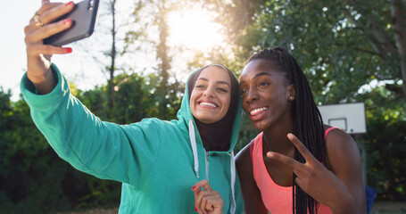 Fototapeta premium Portrait of Two Multiethnic Female Friends in Sports Clothes Posing for a Selfie Using a Smartphone. Young Women Celebrating a Win on an Outdoor Basketball Court by Taking a Photo for Social Media