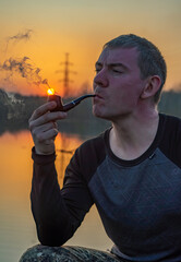 Fototapeta premium Young man smoking a pipe on the bank of the river at sunset