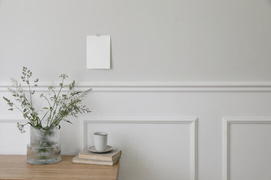 Elegant Scandinavian living room interior. Cup of coffee, books on wooden desk, table. Vase with green grasses and cow parsley. Blank greeting card, invitation mockup taped on wall. White moulding.