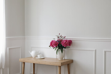 Fototapeta na wymiar Living room still life. Vase with pink peonies flowers. Cup of tea, coffee and tea pot on wooden table near window with curtains. Romantic breakfast. Empty white wall background with elegant moulding.