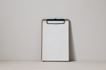 Wooden clipboard mockup with black clip and blank paper sheet on a wooden table against neutral beige wall background. Home office, creative desk. Elegant minimal interior, home decor. Nobody.