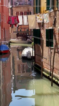 4K vertical video of a narrow Venetian streets with parked boats on green water waves and hinged drying linen across the canal in summer Venice, Veneto region, Italy.