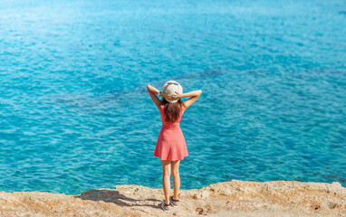 Young woman in coral dress and white hat stands on background of blue sea and sky, back view.