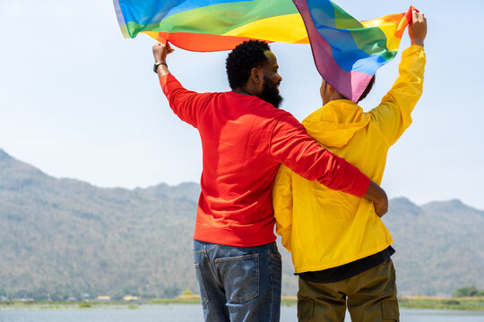 back of gay couple diverse friends standing and waving rainbow flags for celebrating equality sexual freedom, LGBT people enjoy outdoor activities at nature park during summer day in pride month - Powered by Adobe