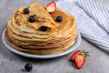 Large stack of traditional thin Russian pancakes on a plate close-up, delicious thin pancakes with berries on a gray concrete table