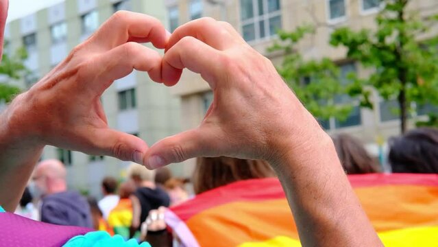 close-up of male hands in heart form against participants of international LGBT movement, Gay pride parade in city with rainbow flags, Pride Month, mass march of lesbian, gay people