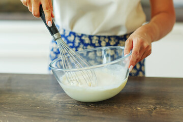 Beautiful hands stir the dough with a black whisk in a deep transparent bowl. 