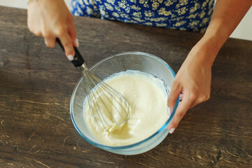 Beautiful hands stir the dough with a black whisk in a deep transparent bowl. 